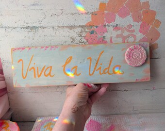 May include: A rectangular wooden sign painted in light blue with the text "Viva la Vida" written in orange script. A small, circular, textured pink medallion featuring an Om symbol is attached to the upper right corner of the wooden board.