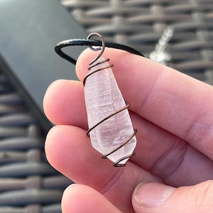 May include: A clear quartz crystal pendant wrapped in copper wire, suspended from a black cord. The crystal is a pale pink hue and has a pointed shape. The pendant is displayed against a dark background.