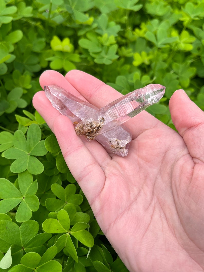 May include: Two clear, pointed crystals with a light purple hue. The crystals are crossed over each other and have a slight shimmer.