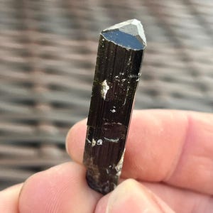 May include: A dark, elongated black tourmaline crystal with a faceted, triangular tip. The crystal has a polished, reflective surface with small, light-colored inclusions. The overall shape is a slender, vertical prism.