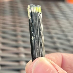 May include: A close-up of a black tourmaline crystal with a rectangular shape. The top of the crystal has a yellow-green hue. The crystal is held by a person's fingers.