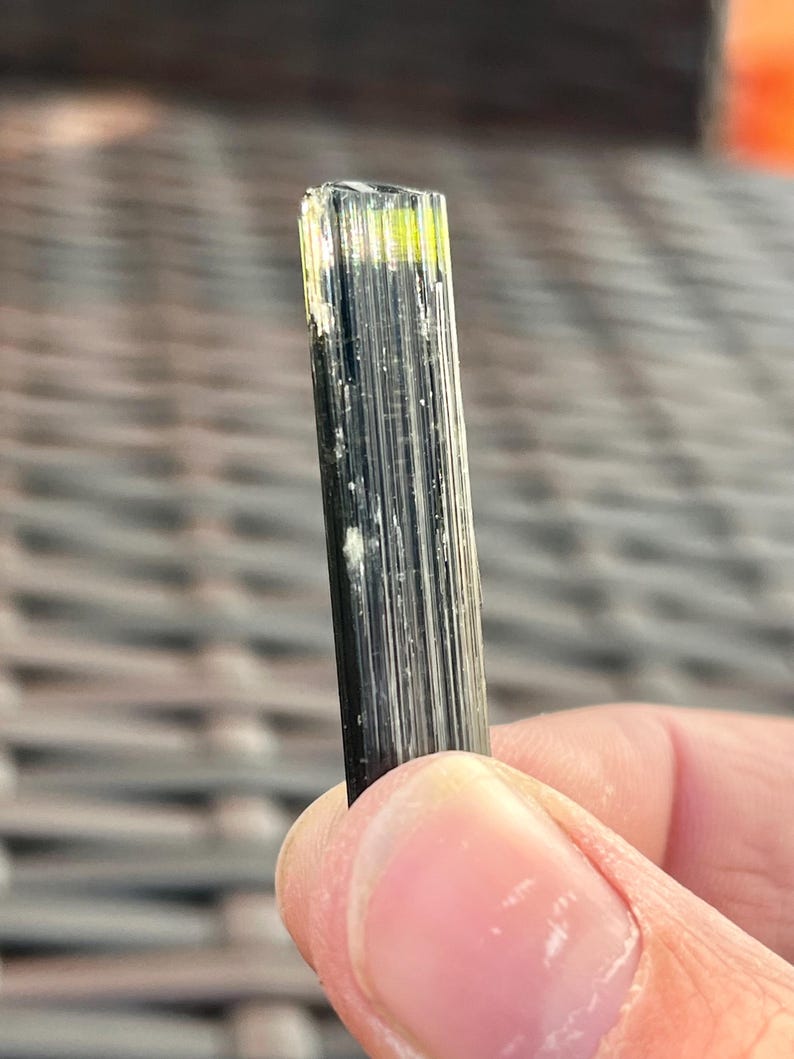 May include: A close-up of a black tourmaline crystal with a rectangular shape. The top of the crystal has a yellow-green hue. The crystal is held by a person's fingers.