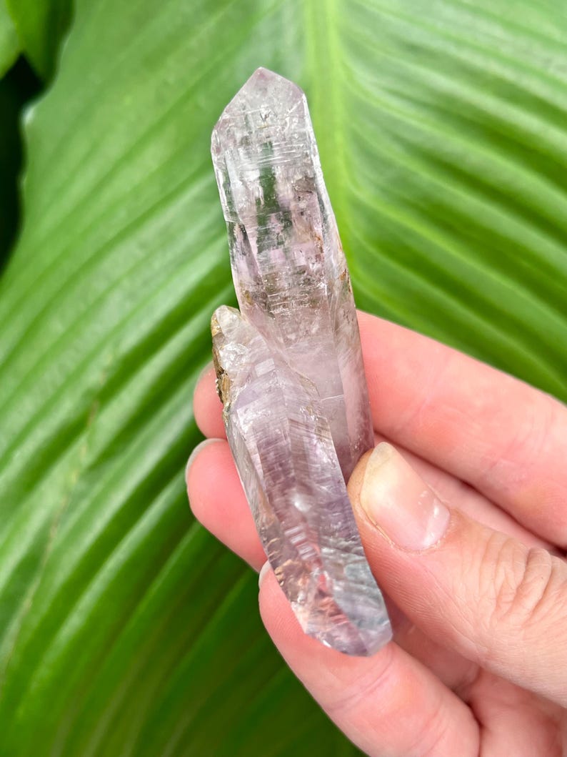 May include: Two amethyst crystal points with a light purple hue and a clear, glassy texture. The crystals are pointed and have a natural, raw appearance.