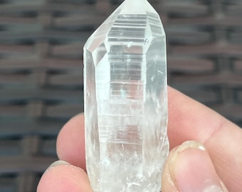 May include: A clear quartz crystal point, held in a hand. The crystal is transparent with visible internal structures and facets. The tip is a lighter shade of white, and the base is rougher in texture.