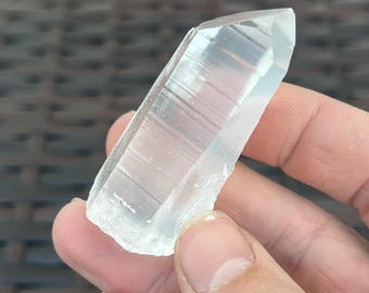 May include: A clear quartz crystal with a pointed tip and a rough, textured base. The crystal is held in a hand, showcasing its translucent quality and natural form. The crystal is approximately 4 inches long.