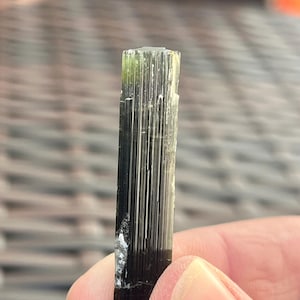 May include: A dark green and black tourmaline crystal with vertical striations. The crystal has a rectangular shape and a slightly rough texture. The top of the crystal is a lighter green color.