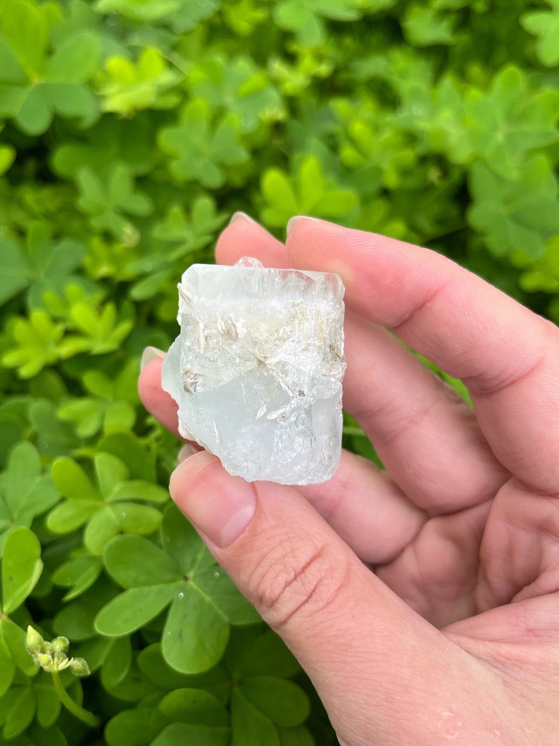 May include: A clear, light blue aquamarine crystal with a rough, natural texture. The crystal has a rectangular shape and is approximately 2 inches in length.