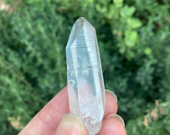 May include: A clear quartz crystal point with a natural, raw finish. The crystal is approximately 2 inches long and has a pointed tip. The crystal is held in a hand against a blurred background of green foliage.