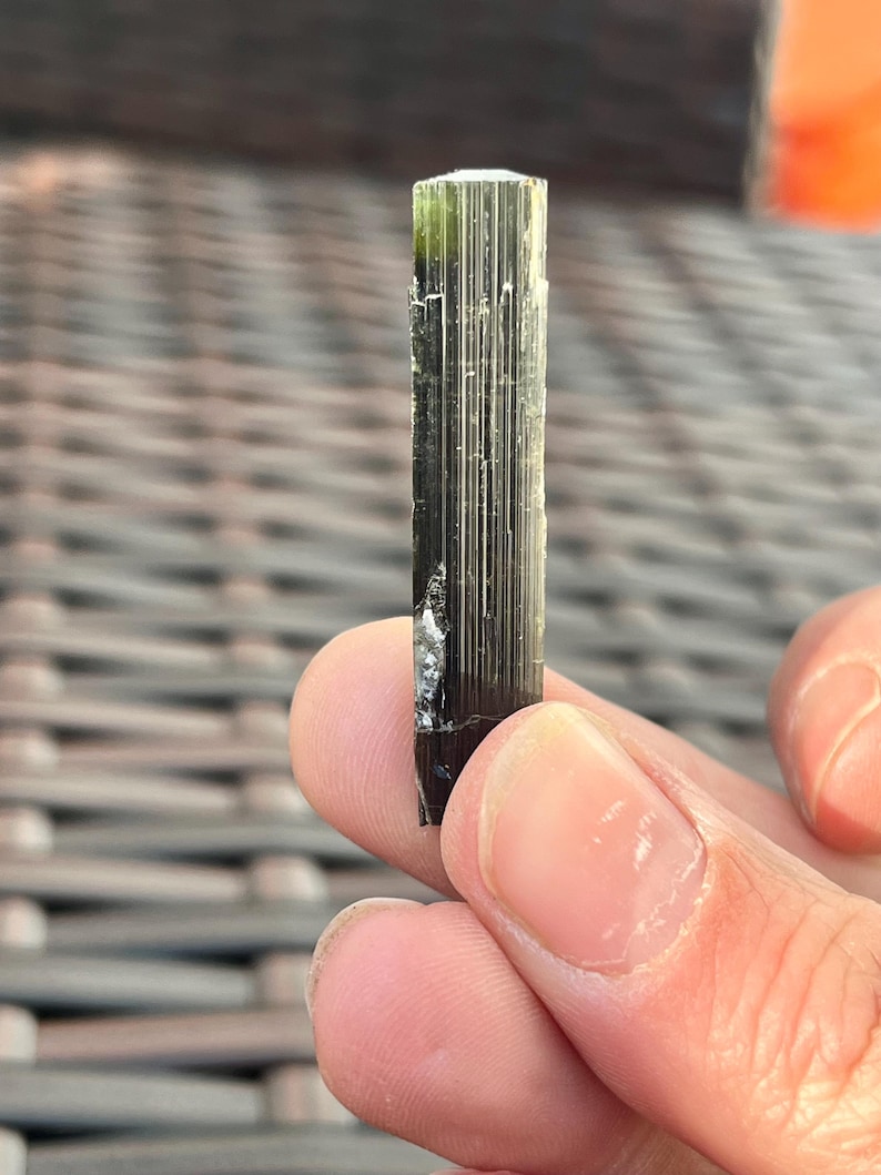 May include: A dark green and black tourmaline crystal with a vertical, columnar structure. The crystal has a translucent, glassy appearance with a slightly rough texture. The top of the crystal is a lighter green color.