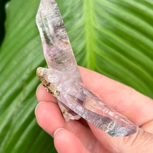 May include: A double-terminated amethyst crystal with a clear, light purple hue. The crystal has a natural, raw appearance with a slight green tint at the tip.