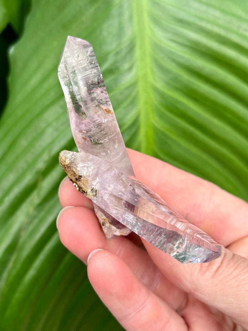 May include: A double-terminated amethyst crystal with a clear, light purple hue. The crystal has a natural, raw appearance with a slight green tint at the tip.