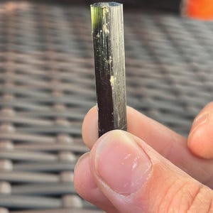 May include: A dark green and black tourmaline crystal with a columnar structure. The gemstone is held in a hand, showcasing its natural, raw form. The crystal's surface has vertical striations and a slight translucence.
