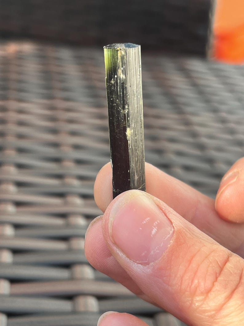 May include: A dark green and black tourmaline crystal with a columnar structure. The gemstone is held in a hand, showcasing its natural, raw form. The crystal's surface has vertical striations and a slight translucence.
