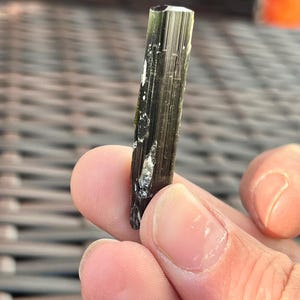 May include: A dark green tourmaline crystal with a faceted top. The crystal is held between fingers and has white inclusions and vertical striations. The crystal is approximately 1 inch in length.