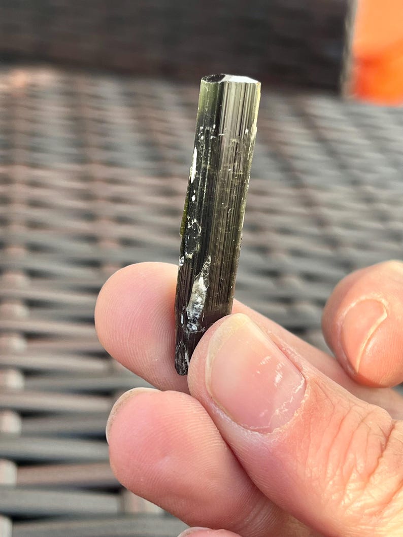 May include: A dark green tourmaline crystal with a faceted top. The crystal is held between fingers and has white inclusions and vertical striations. The crystal is approximately 1 inch in length.