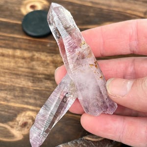 May include: Two clear quartz crystal points with a light purple hue. The crystals are pointed at the top and have a natural, rough texture.