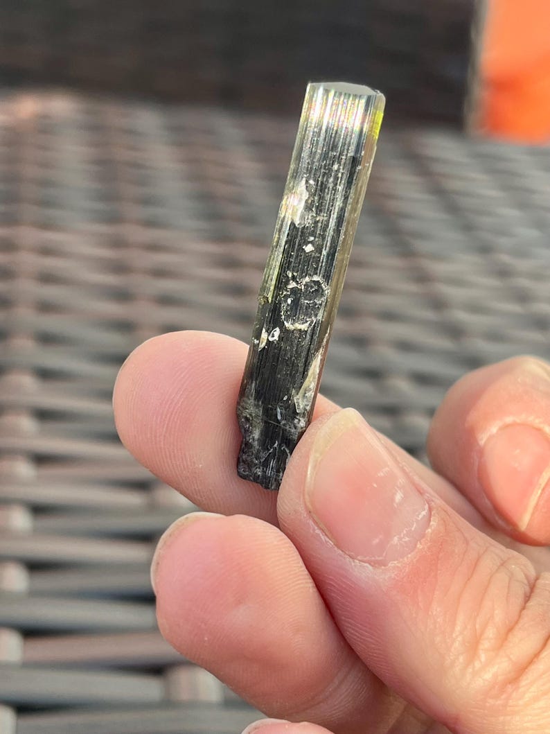 May include: A raw, elongated tourmaline crystal with a dark green and black color. The crystal is held between fingers, showcasing its natural, textured surface and subtle color variations. The top of the crystal has a yellow-green hue.