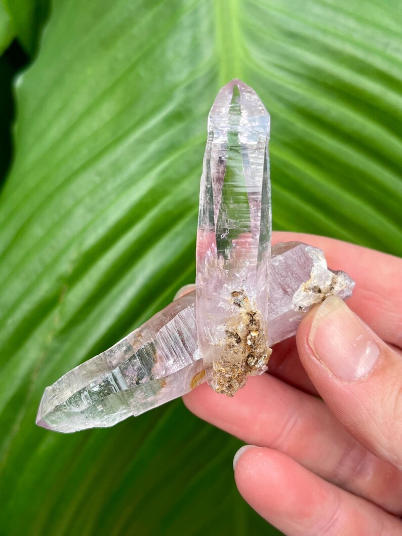 May include: Two clear quartz crystals with a purple hue, one is larger than the other, and both have a natural, raw look.