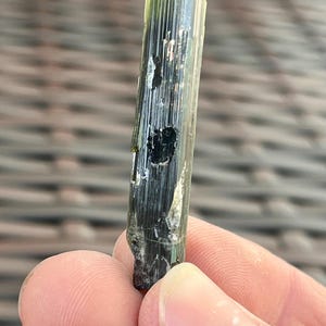 May include: A raw, elongated tourmaline crystal with a dark green hue. The crystal has a vertical, striated texture and a slightly irregular shape. The top of the crystal is a darker shade of green, and the bottom is a rough, uneven surface.