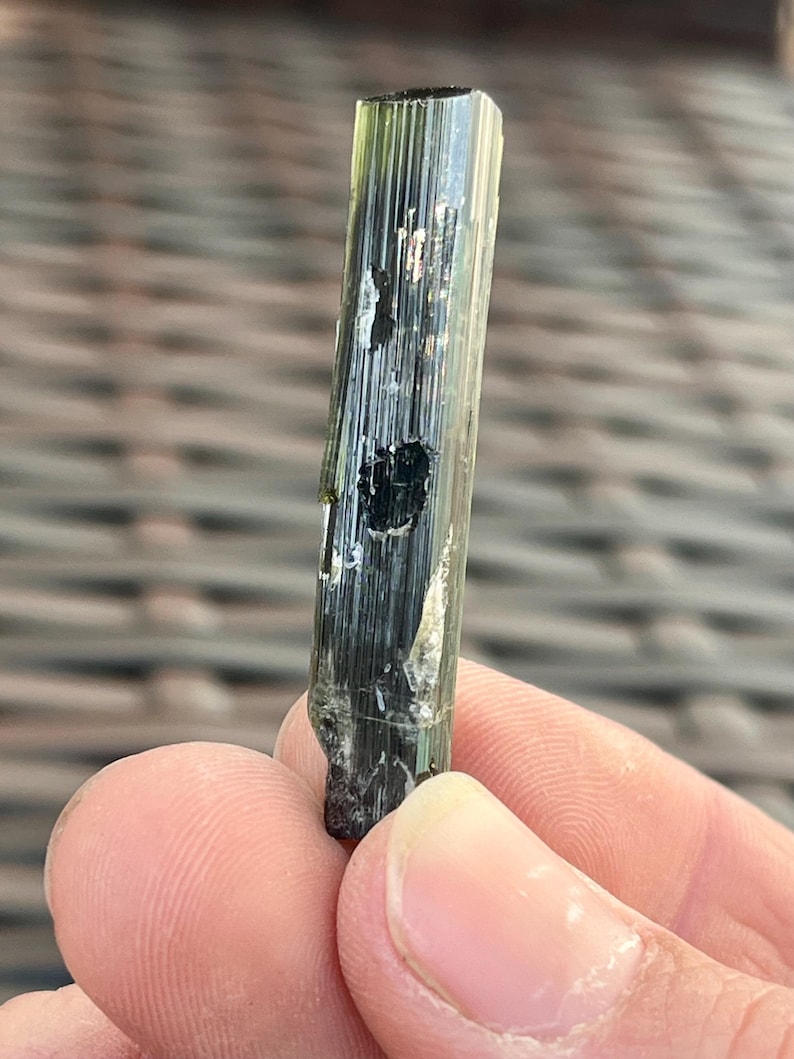 May include: A raw, elongated tourmaline crystal with a dark green hue. The crystal has a vertical, striated texture and a slightly irregular shape. The top of the crystal is a darker shade of green, and the bottom is a rough, uneven surface.