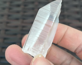 May include: A clear, crystalline quartz point with a textured, striated surface. The quartz is held in a hand, showcasing its natural, raw form. The crystal's facets catch the light, highlighting its translucent quality. This is a natural gemstone.