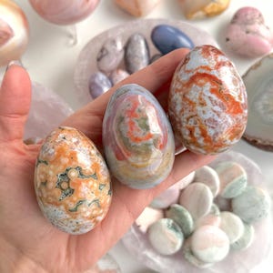 Puede incluir: Una mano sostiene tres huevos de piedra pulida con patrones naranjas, blancos y grises. En el fondo se ven otras piedras pulidas de varias formas y colores, incluyendo corazones y esferas. Los huevos miden aproximadamente 7,6 cm de altura.