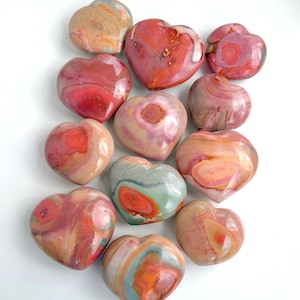 Può includere: Una collezione di pietre a forma di cuore in vari colori, tra cui rosso, rosa, arancione e grigio. Le pietre lucide sono disposte su uno sfondo bianco. Le pietre sembrano essere fatte di un materiale naturale, forse una gemma.