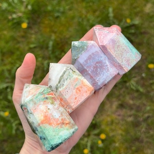 May include: Four colorful gemstone towers are held in a hand. The crystals have a variety of colors, including green, orange, pink, and purple. The crystals are faceted and have a polished appearance. The background is blurred green grass.