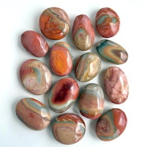 Può includere: Un assortimento di pietre levigate di varie forme e dimensioni. Le pietre presentano una gamma di colori, tra cui rosso, arancione, marrone e blu, con motivi e vortici unici. Le pietre sono disposte su una superficie bianca.