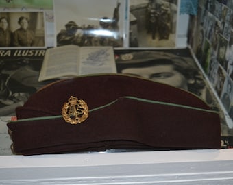 Może przedstawiać: Bordowa wojskowa czapka z złotym emblematem z literami "ATS" i koroną. Czapka ma jasnozielone wykończenie wzdłuż górnej krawędzi. Tło obejmuje zabytkowe zdjęcia i magazyn.