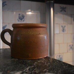 Op de afbeelding: Een bruine keramische pot met een handvat. De pot heeft een afgeronde body en een licht uitlopende rand. Hij staat op een donker aanrecht, met een betegelde muur op de achtergrond. De tegels hebben blauwe bloempatronen.