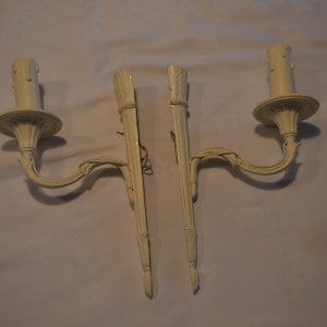 Può includere: Due applique da parete bianco sporco con un design a candela. Ogni applique presenta un braccio curvo con un elemento decorativo, un portacandele e una lunga piastra posteriore. Le applique sono realizzate in materiale chiaro.