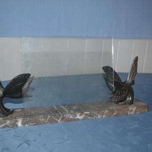 Può includere: Un fermalibri decorativo in vetro trasparente e marmo con due sculture di uccelli stilizzati. Gli uccelli sono grigio scuro con ali dettagliate, montati su una base rettangolare in marmo venato marrone e bianco.