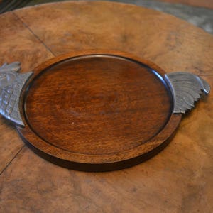 Peut inclure: Un plateau de service rond en bois avec une finition foncée. Le plateau a un bord surélevé et est orné de poignées en forme de tête de coq argentées. Le plateau est posé sur une table en bois.