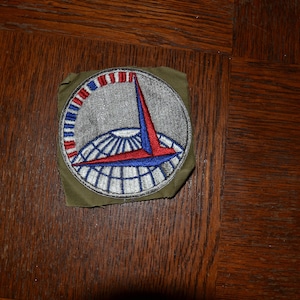 Op de afbeelding: Een vintage militaire patch met een zilver, blauw en rood ontwerp. De patch toont een gestileerd vliegtuig met een wereldbol en een rood-wit-blauwe streep.