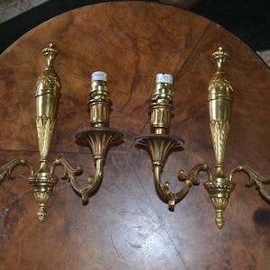 Può includere: Coppia di applique antiche in ottone. Ogni applique presenta un design ornato con due portalampada. Le applique sono montate su una superficie in legno, evidenziando il loro stile elegante e vintage.