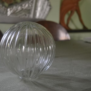 Op de afbeelding: Een bolvormige lampenkap van helder glas met een verticale ribbelstructuur. De bol is transparant met een gladde, ronde bovenkant en een cirkelvormige opening aan de onderkant, ontworpen om op een lamphouder te worden bevestigd.