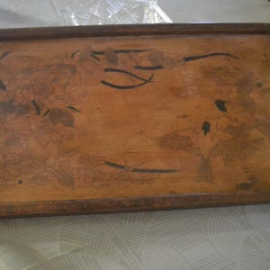 Peut inclure: Un plateau de service rectangulaire en bois avec une finition brun foncé et deux poignées latérales verticales. La surface présente un motif détaillé de fleurs épanouies sur la gauche et de deux oiseaux en vol sur la droite.