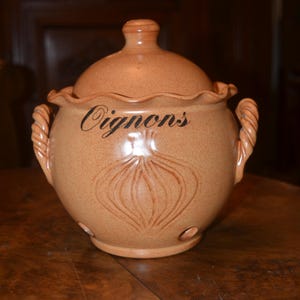 May include: A tan ceramic onion keeper with a lid and rope-style handles. The word "Oignons" is written in black script on the front, above an embossed onion design. The keeper has small holes near the base.