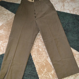 May include: Brown trousers with a button fly closure. The pants have a high waist and a straight leg design. The fabric appears to be a durable, medium-weight material. The trousers are laid flat on a patterned rug.