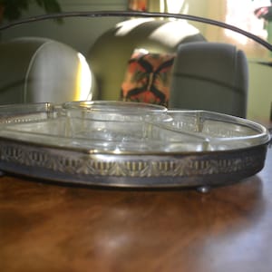 Könnte beinhalten: Eine ovale Glasschale mit vier unterteilten Fächern, eingefasst in einen dekorativen silberfarbenen Metallrahmen mit einem hohen, gebogenen Griff und kleinen kugelförmigen Füßen. Die klaren Glasabschnitte weisen ein strukturiertes Muster auf.