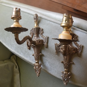Può includere: Un paio di applique da parete dorate ornate con un motivo a volute decorative. Ogni applique ha un'unica presa per lampadina e una base in ceramica bianca.