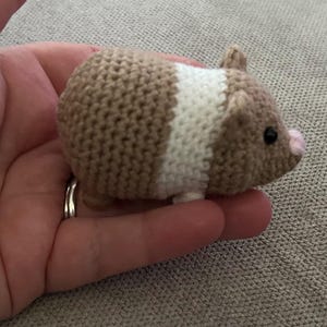 May include: A small, crocheted guinea pig toy featuring a tan and white color-blocked pattern. The plush figure has a rounded body, two small ears, a tiny pink nose, and black bead eyes, crafted with a visible yarn stitch texture throughout.