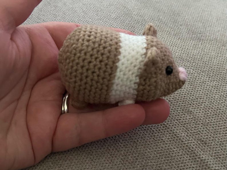 Op de afbeelding: Een klein gehaakt cavia-speeltje met een beige en wit kleurblokpatroon. Het pluche figuurtje heeft een rond lichaam, twee kleine oortjes, een piepklein roze neusje en zwarte kraaloogjes, afgewerkt met een zichtbare gebreide textuur.