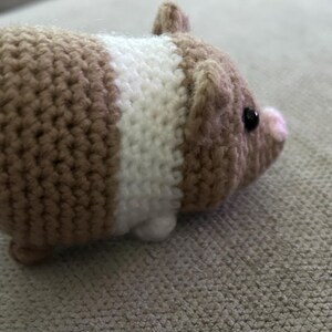 Op de afbeelding: Een kleine, gehaakte cavia-figuur met een rond lichaam van beige en wit garen. Het pluche speelgoed heeft een kleine roze neus, twee piepkleine oortjes en &eacute;&eacute;n zichtbaar zwart kraaloog aan de zijkant van de kop, gemaakt met een textuursteek.