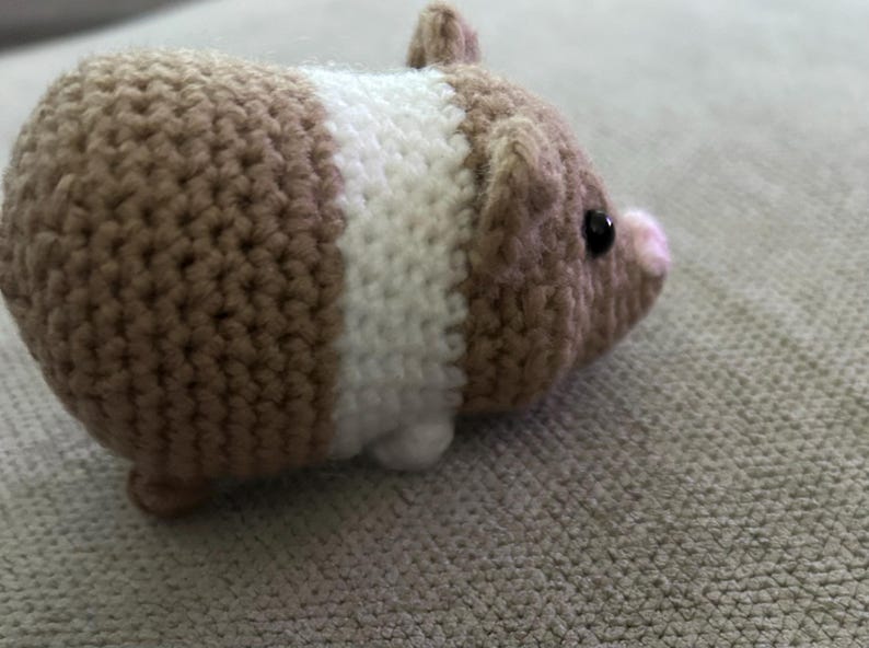 Op de afbeelding: Een kleine, gehaakte cavia-figuur met een rond lichaam van beige en wit garen. Het pluche speelgoed heeft een kleine roze neus, twee piepkleine oortjes en &eacute;&eacute;n zichtbaar zwart kraaloog aan de zijkant van de kop, gemaakt met een textuursteek.