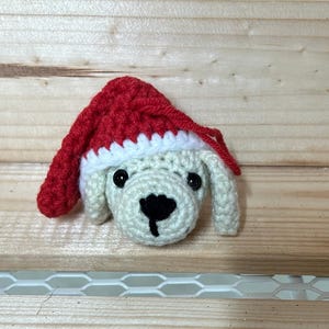 May include: A small crocheted dog wearing a red and white Santa hat. The dog is white with black eyes and a black nose.