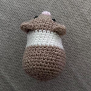 Op de afbeelding: Een klein, rond, gehaakt hamsterspeeltje met een bruine kop en onderkant en een brede witte band in het midden. De hamster heeft twee kleine zwarte kraaloogjes, een piepklein roze neusje en twee afgeronde oortjes boven op de kop.