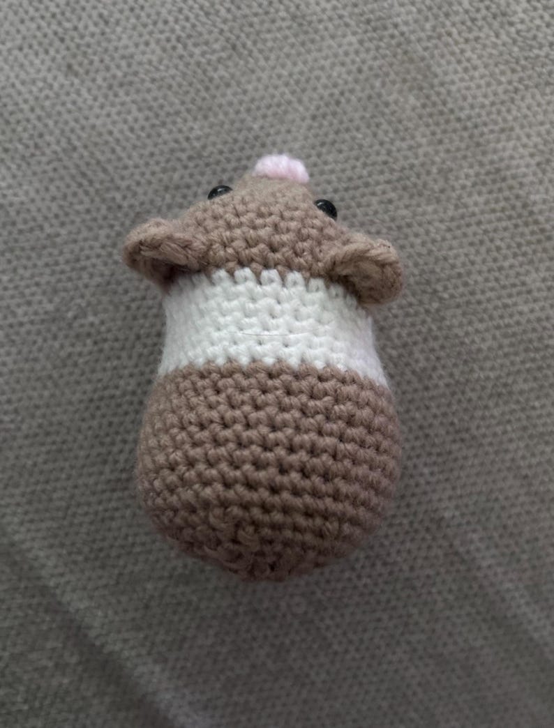 Op de afbeelding: Een klein, rond, gehaakt hamsterspeeltje met een bruine kop en onderkant en een brede witte band in het midden. De hamster heeft twee kleine zwarte kraaloogjes, een piepklein roze neusje en twee afgeronde oortjes boven op de kop.
