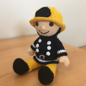Puede incluir: Un juguete de bombero de crochet que lleva un casco amarillo con una visera negra y un uniforme blanco y negro con lunares blancos.